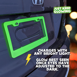 Close-up of a green glow in the dark license plate frame attached to a dark-colored car, highlighting its bright glow and unique design. Text indicates it charges with any light and is best viewed in the dark. Just Keep Glowing.